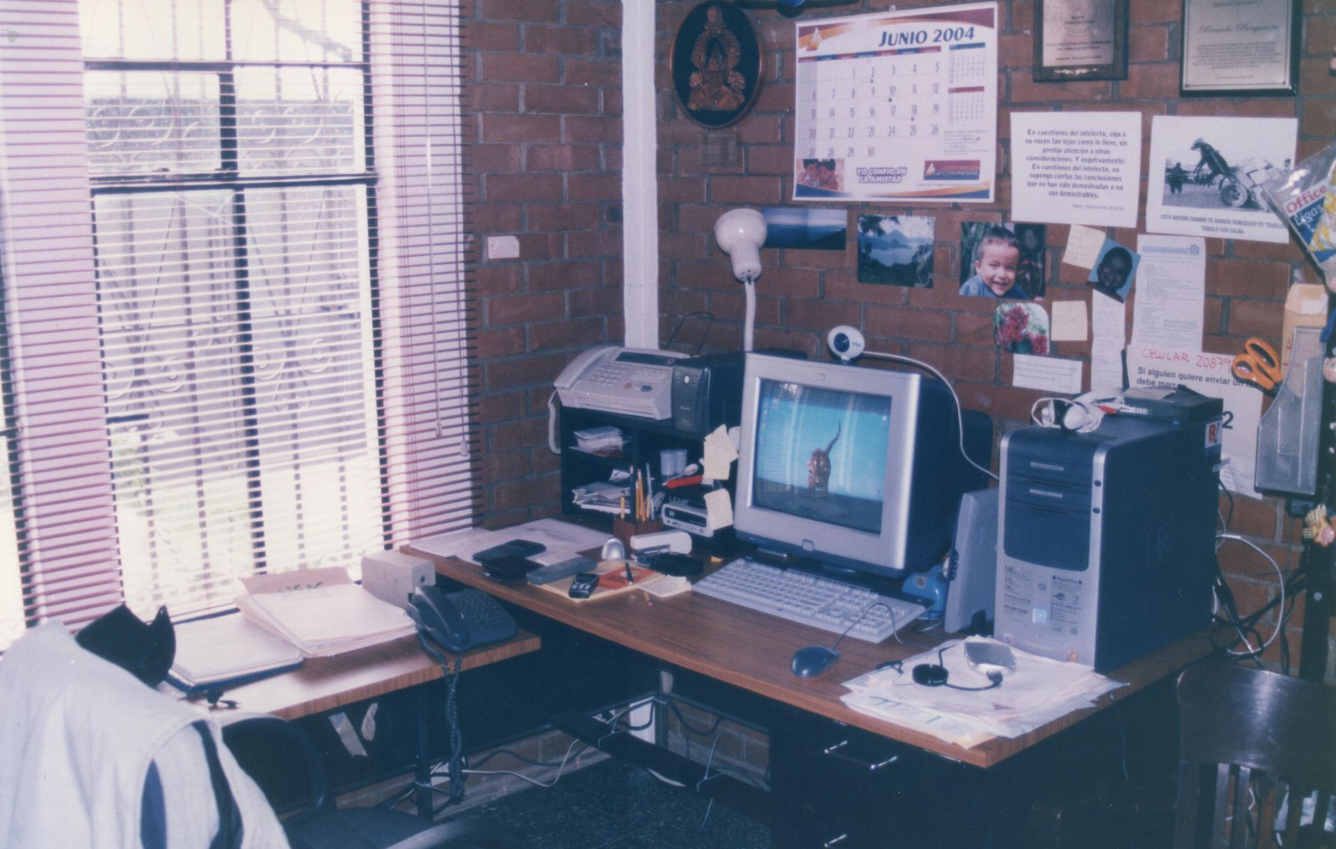 Office desk with computer, printer, and accessories; window with blinds; brick wall.