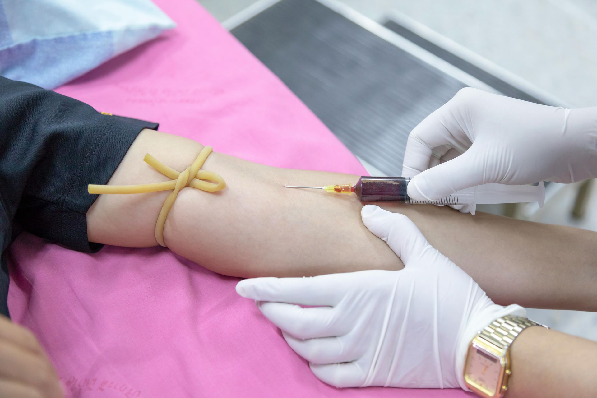 A person's arm with a tourniquet has a needle inserted for blood draw. White-gloved hands are assisting.