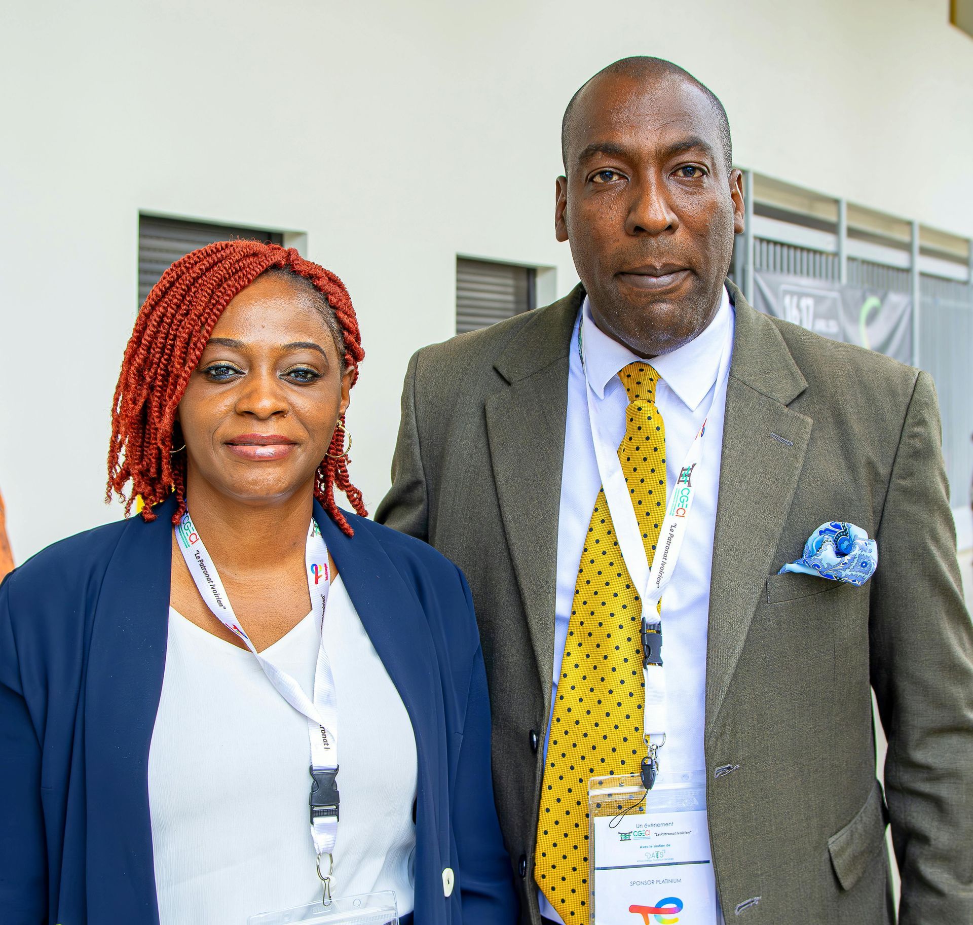 Woman with red dreadlocks and man in a suit stand side by side, both with lanyards.